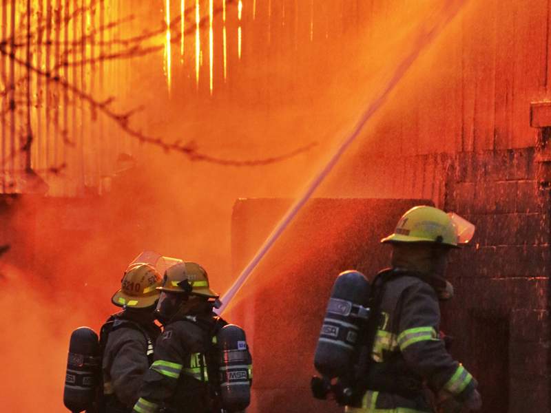 Firefighters direct a high-pressure water hose at a building engulfed in flames and smoke during a fire response operation.
