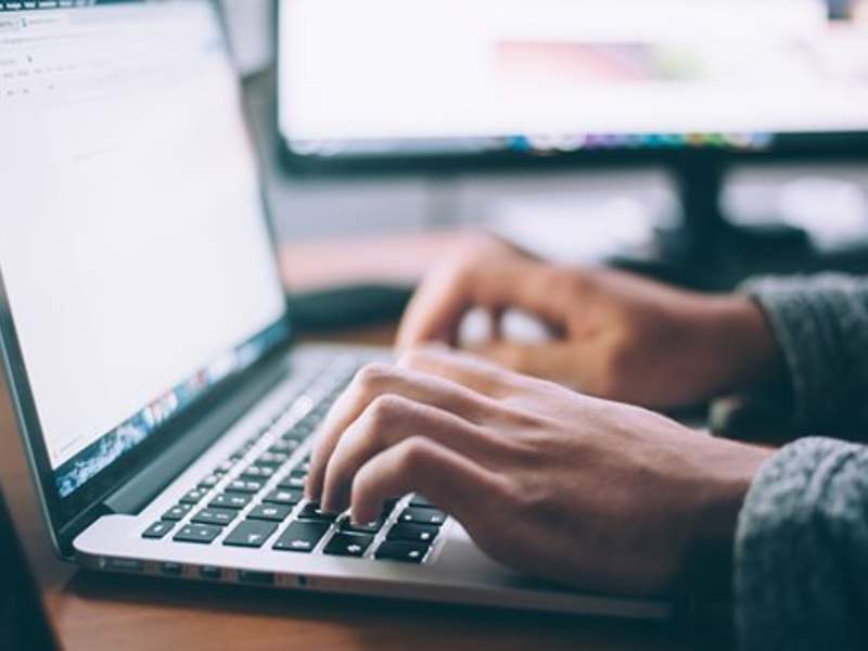Hands typing on a laptop keyboard with a blurred monitor in the background.