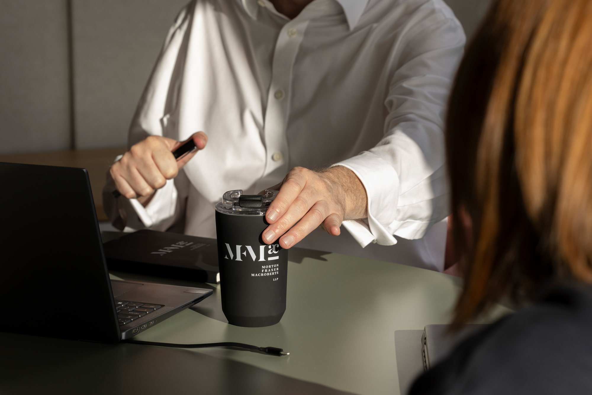 Two people in a meeting with laptops and an MFMac-branded reusable cup on the table.