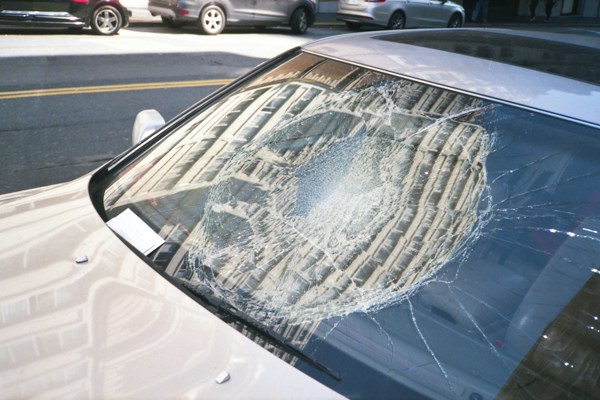 Close-up of a parked car with a heavily cracked windscreen, showing a large circular impact area with radiating fractures; surrounding street and buildings are reflected in the damaged glass.