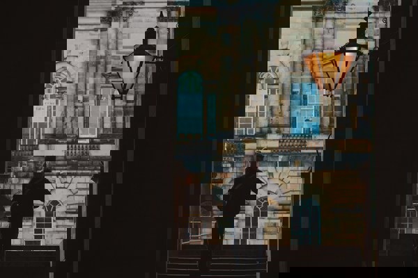 View through an archway showing stone steps, a historic stone building, and a single person walking beneath a traditional street lamp.