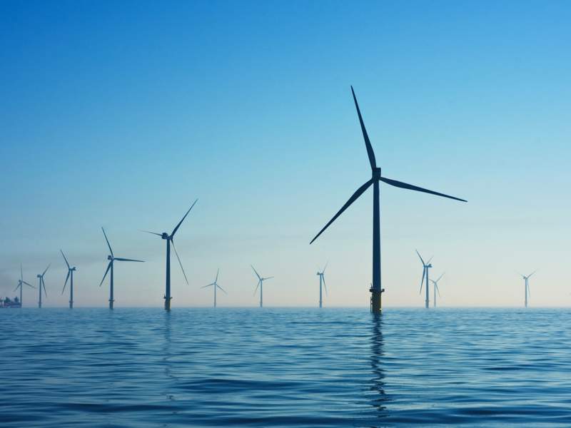 Offshore wind farm with multiple turbines spread across calm ocean waters under a clear blue sky, with a ship visible in the distance on the left.