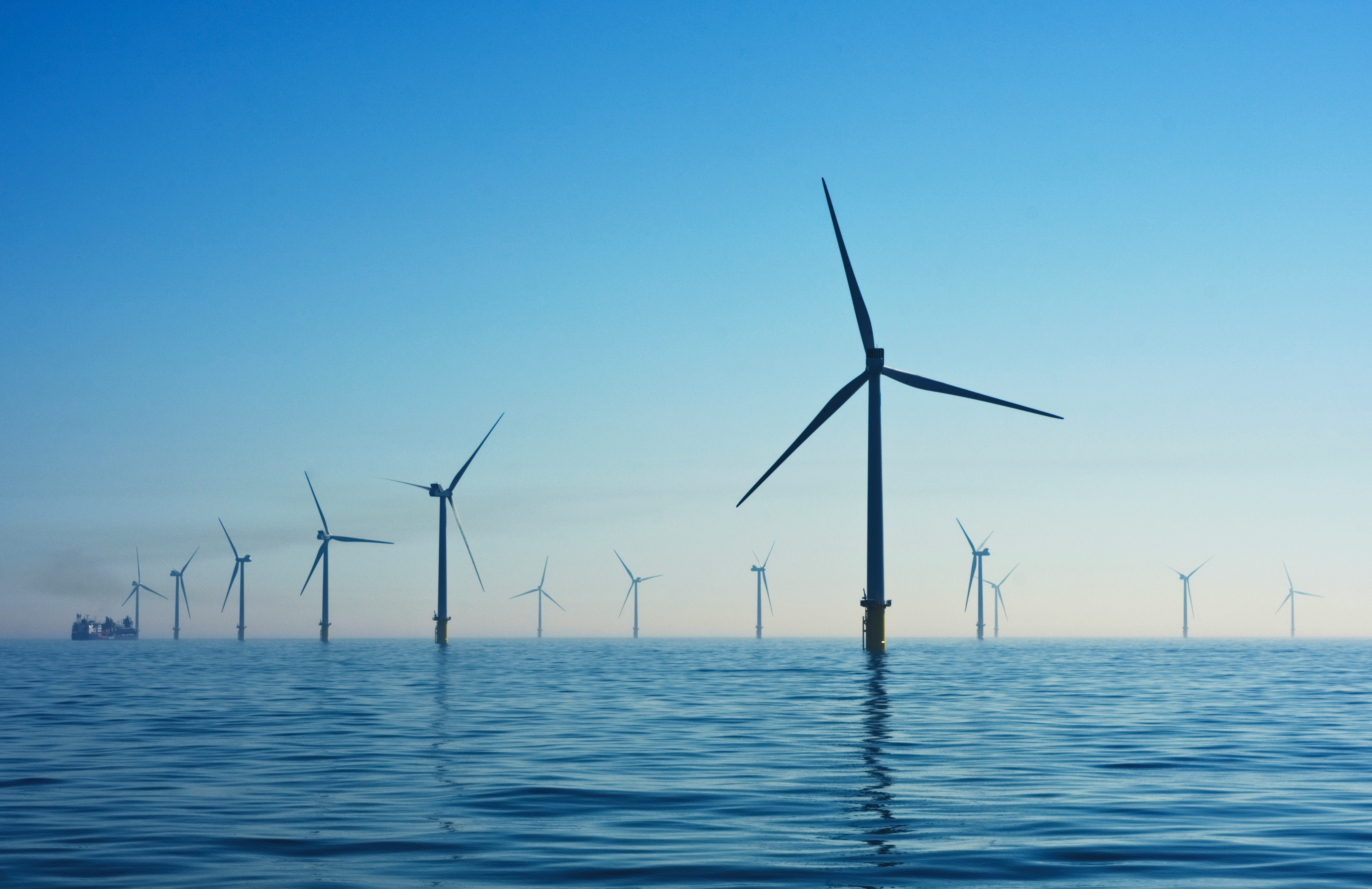 Offshore wind farm with multiple turbines spread across calm ocean waters under a clear blue sky, with a ship visible in the distance on the left.