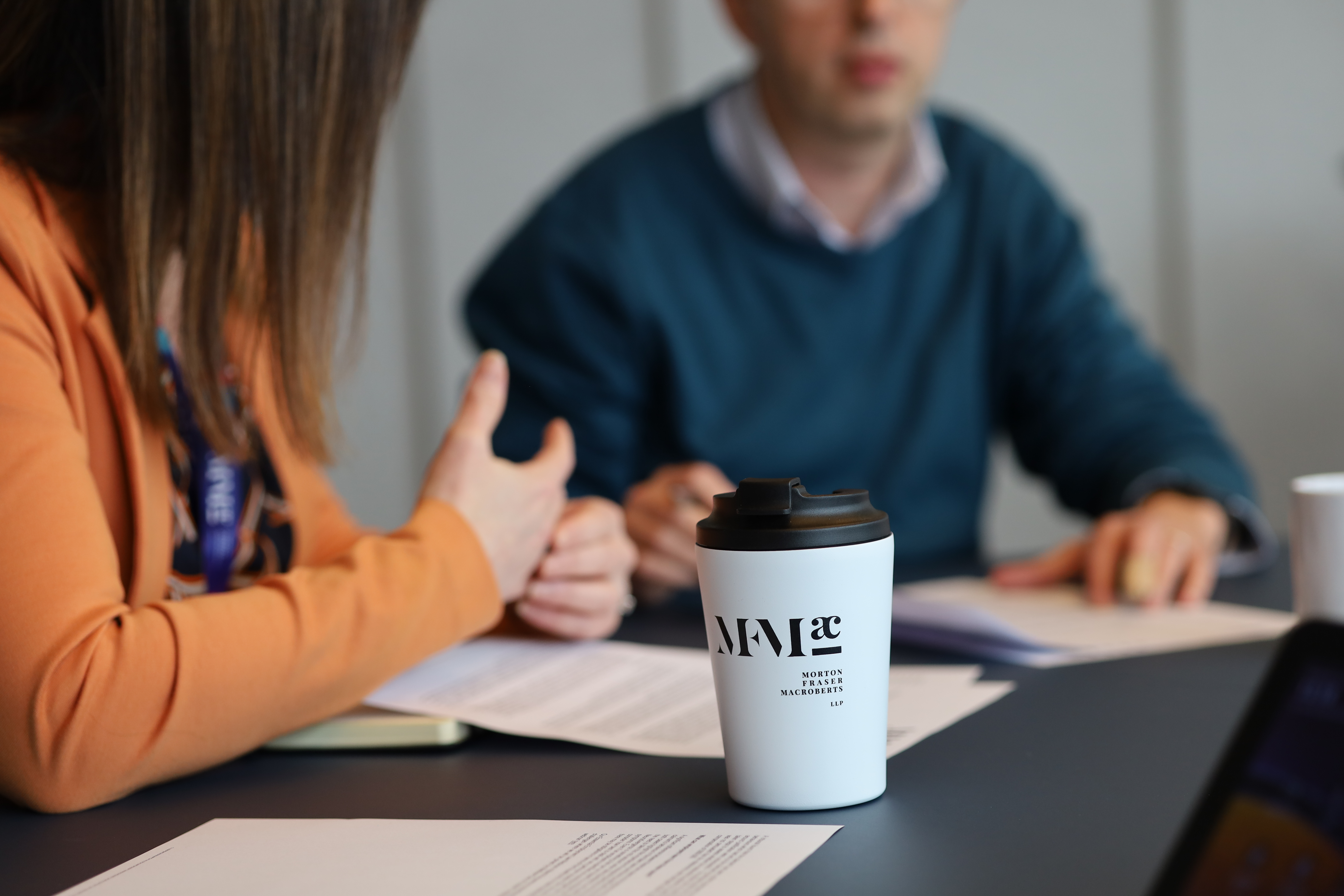 Two people seated at a meeting table reviewing documents, with a branded MFMac takeaway coffee cup in the foreground and paperwork visible on the table.