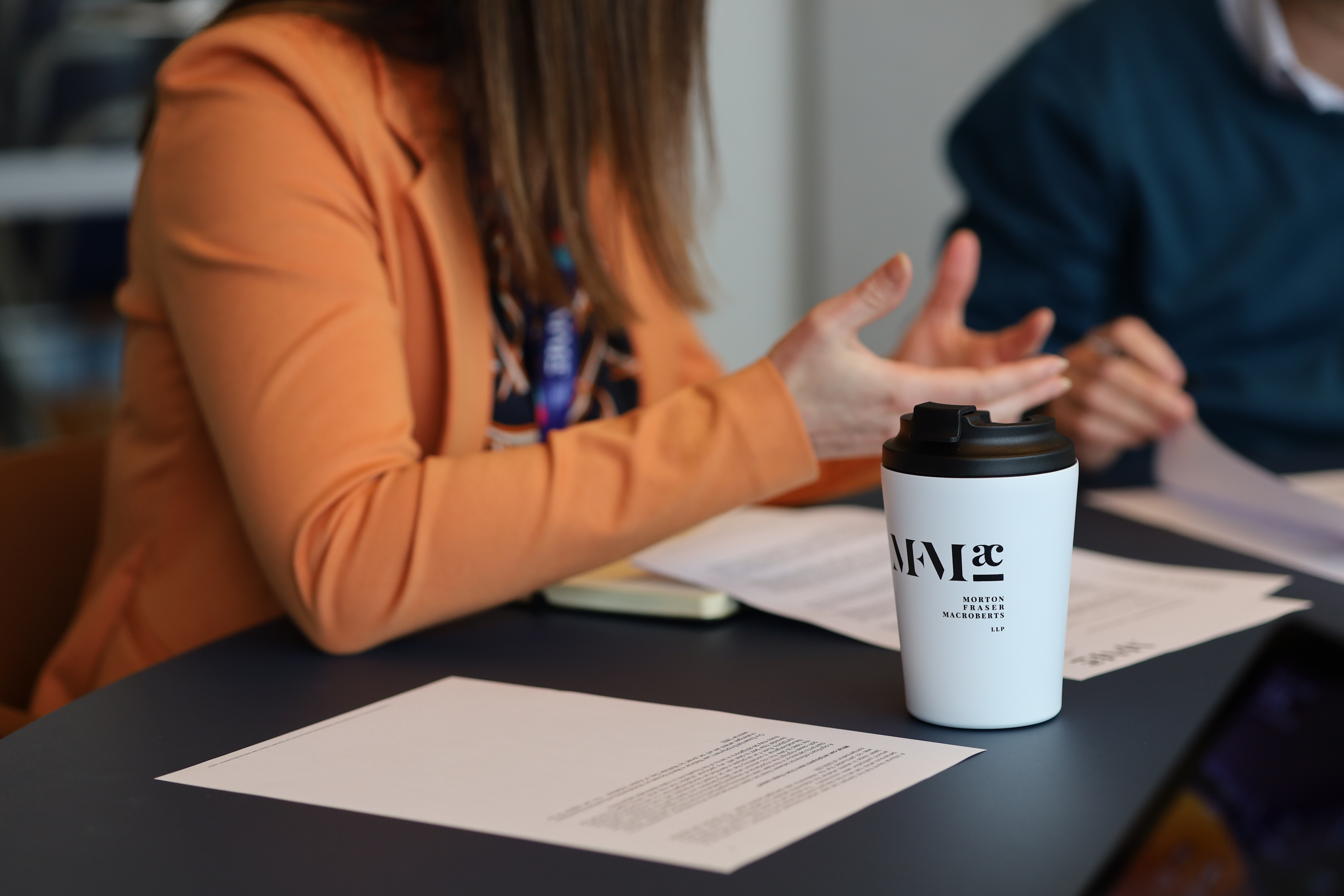 MFMac-branded coffee cup on a meeting table beside documents, with professionals engaged in discussion in the background, illustrating workplace compliance and employment-related matters.