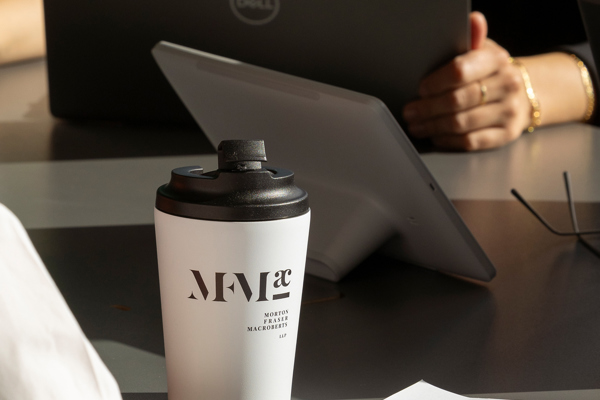 Meeting table with a laptop, tablet and printed draft document, with an MFMac branded coffee cup in the foreground.