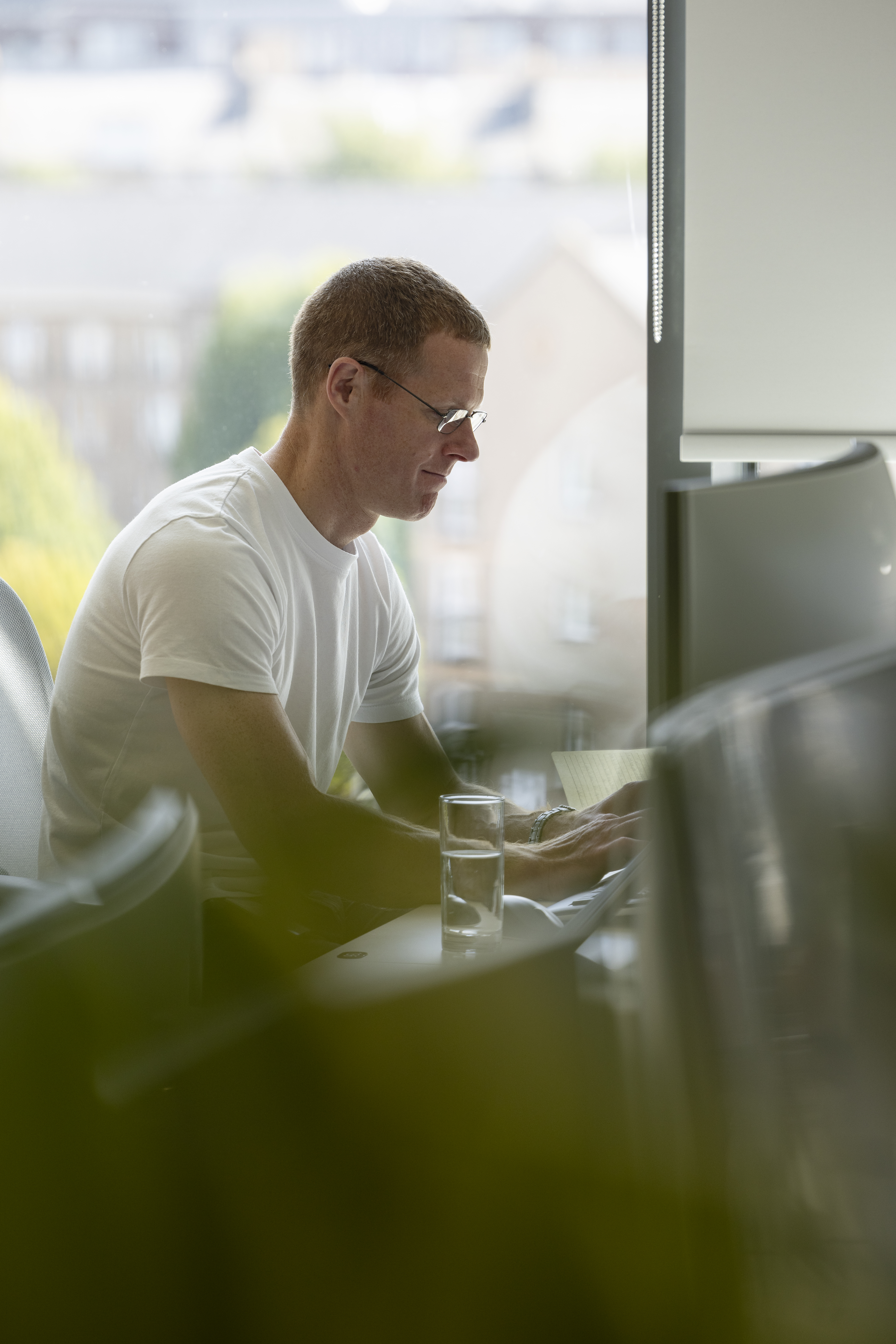 Person wearing glasses working on a laptop at a desk in a bright office, with a glass of water in the foreground and a large window in the background.