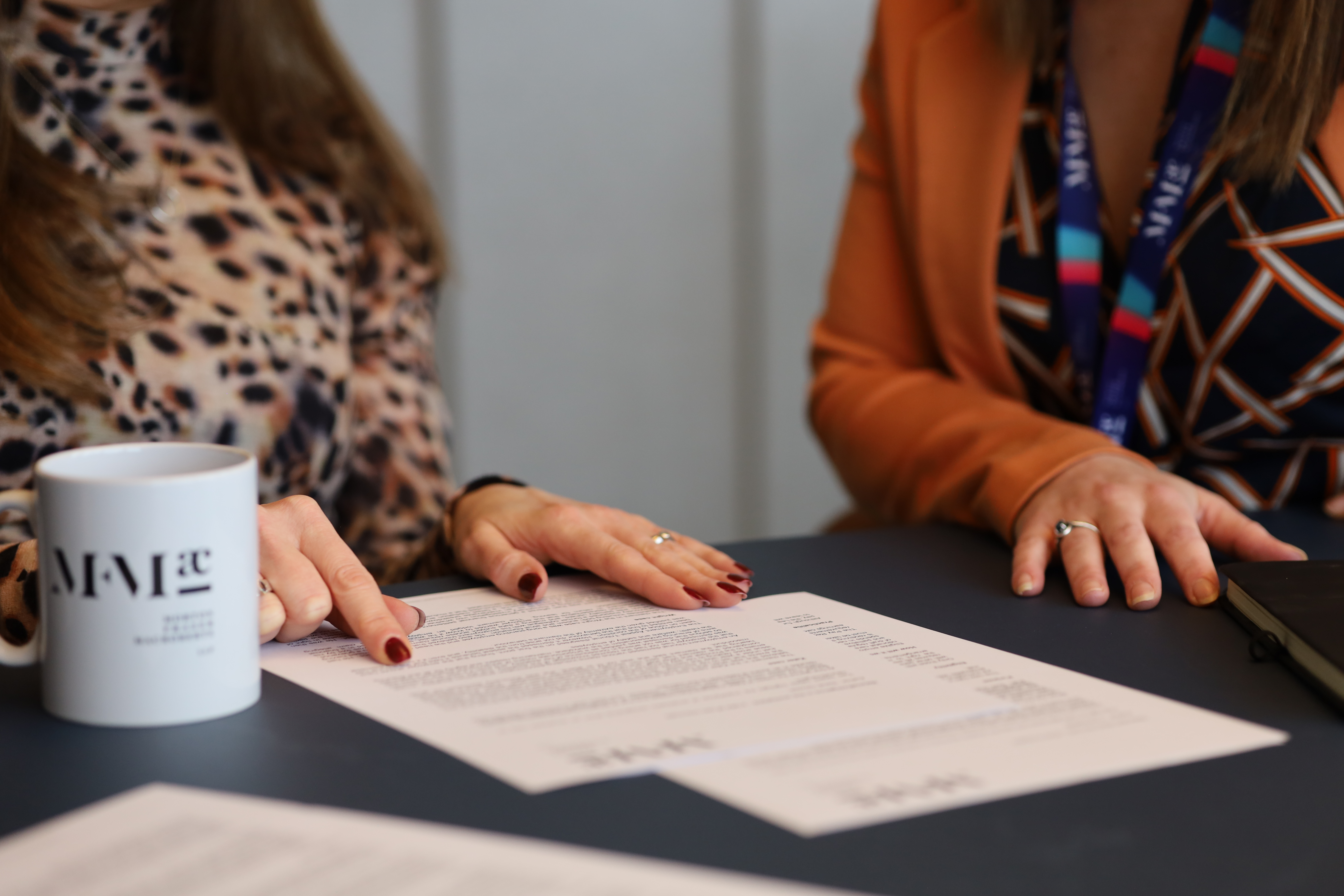 Two colleagues review a document at a meeting table, with paperwork, a notebook and an MFMac-branded mug visible in the foreground.