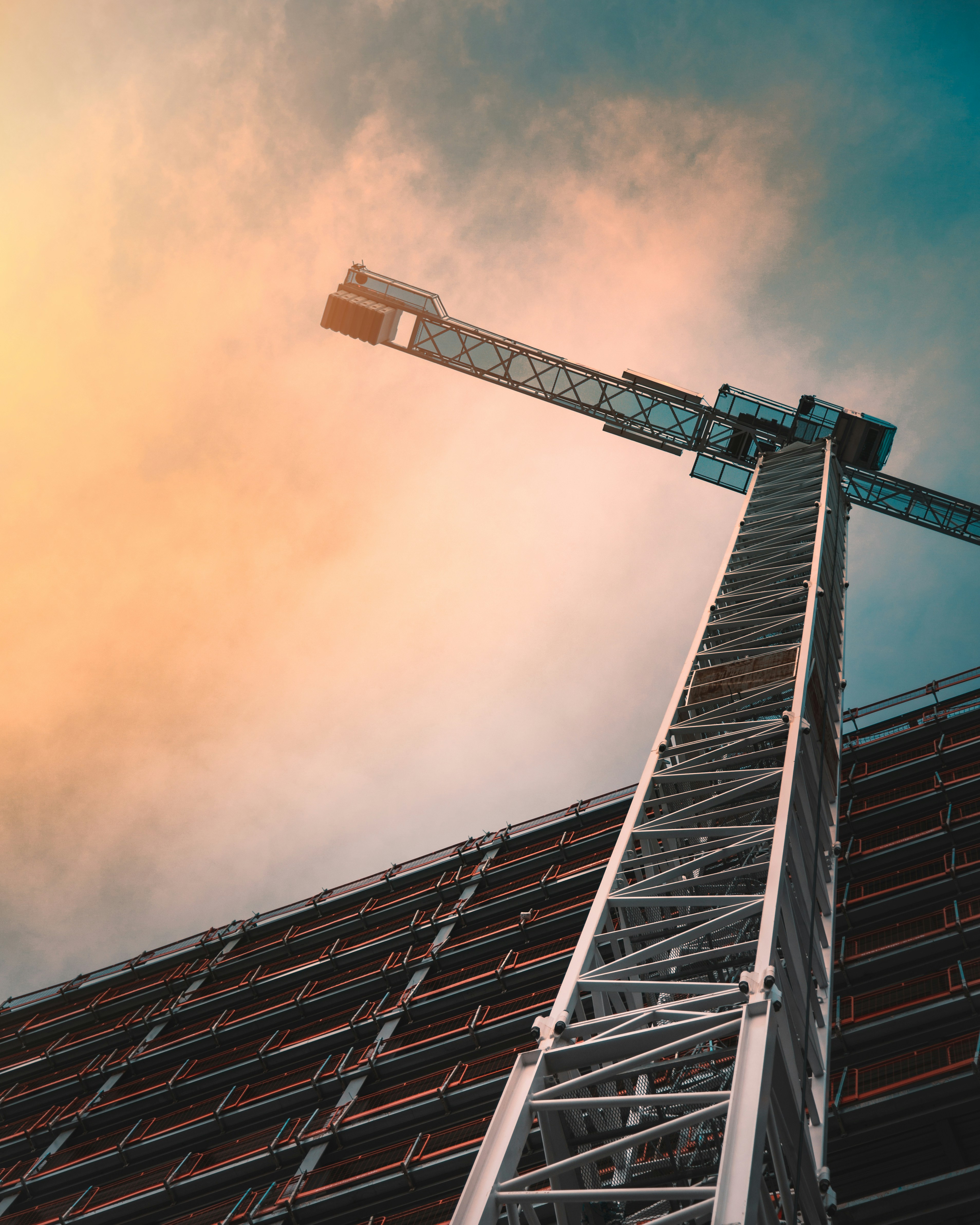 Low-angle view of a tower crane beside a high-rise building under construction, with the crane's structure and the building façade silhouetted against a dramatic sky.