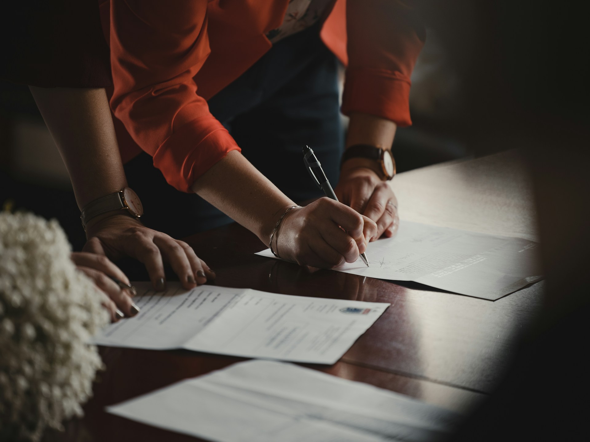 Person signing a document on a table while others review related paperwork, illustrating formal agreement or legal documentation.