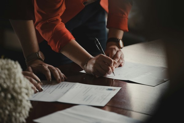 Person signing a document on a table while others review related paperwork, illustrating formal agreement or legal documentation.