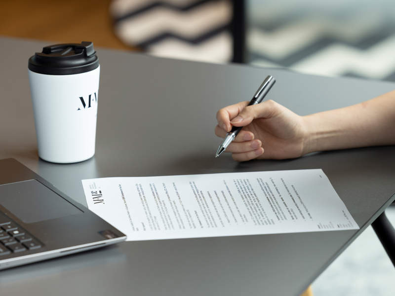 Hand holding a pen over a printed contract document on a desk, with a laptop and takeaway coffee cup beside it.