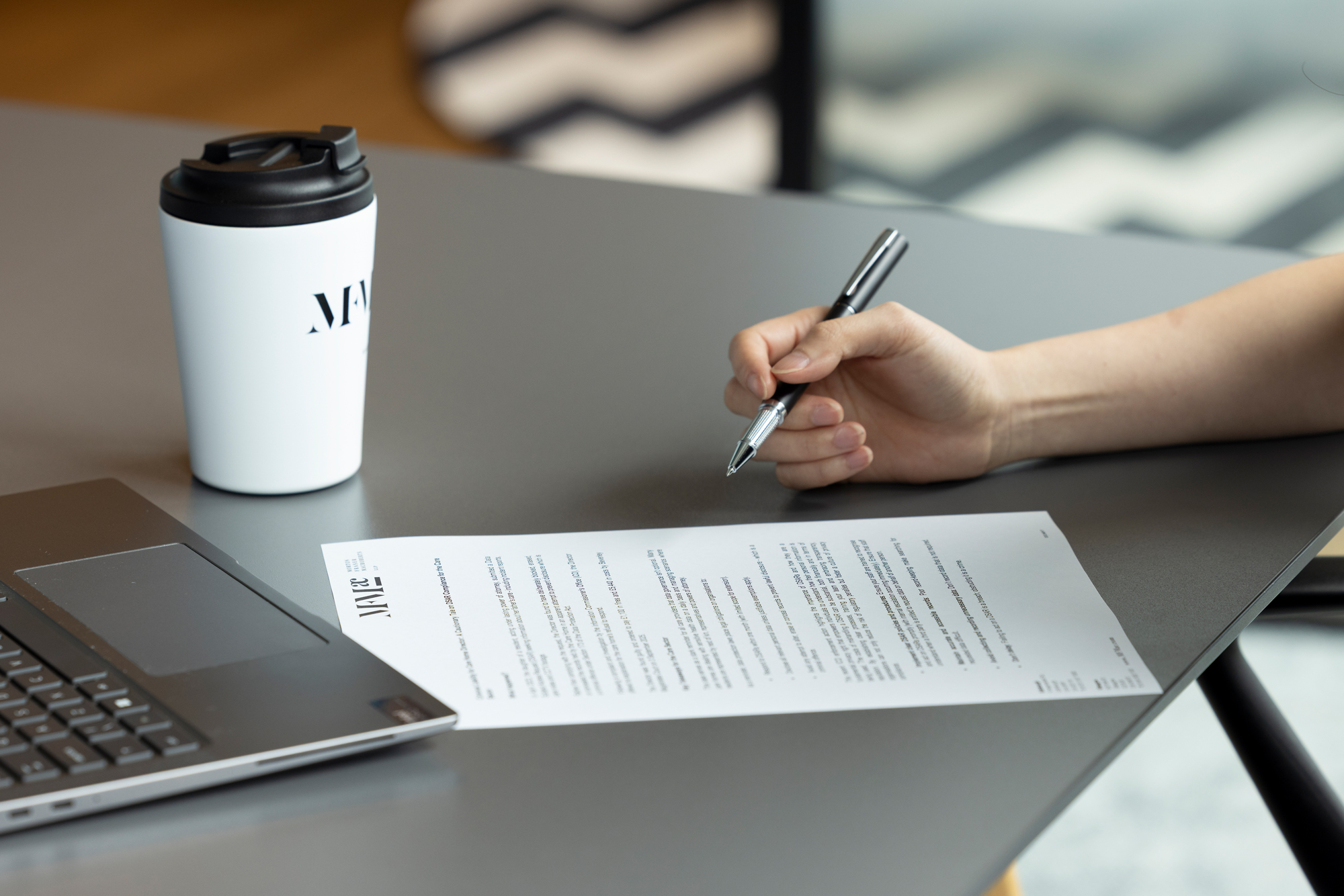 Hand holding a pen over a printed contract document on a desk, with a laptop and takeaway coffee cup beside it.