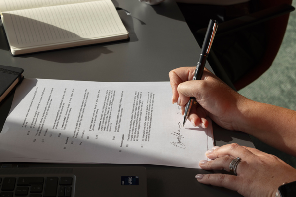 Close-up of a person signing a formal document on a desk beside a laptop and notebook, representing the UK visa application process
