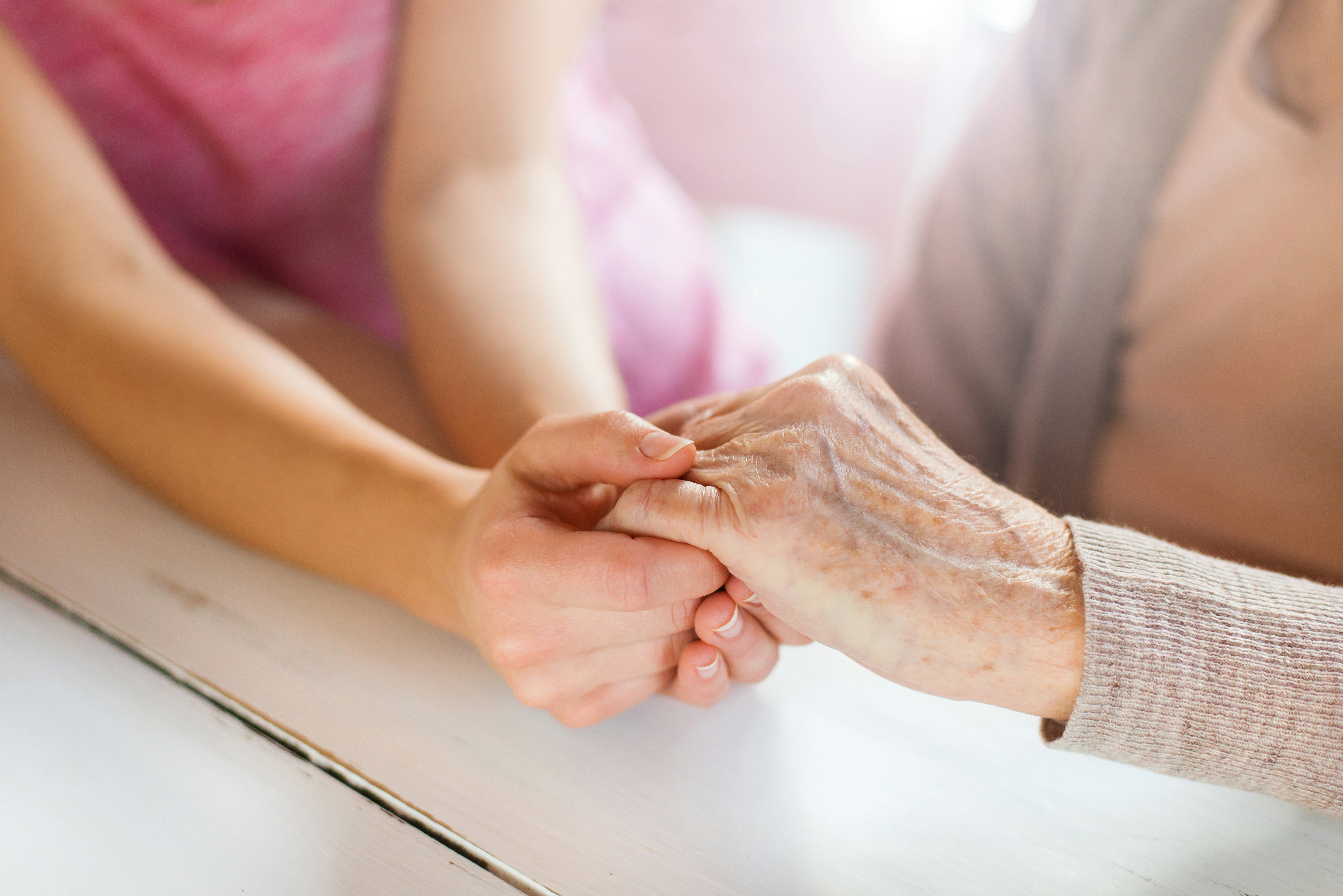 Close-up of a younger person gently holding an older person's hand across a table, symbolising support, care and companionship.