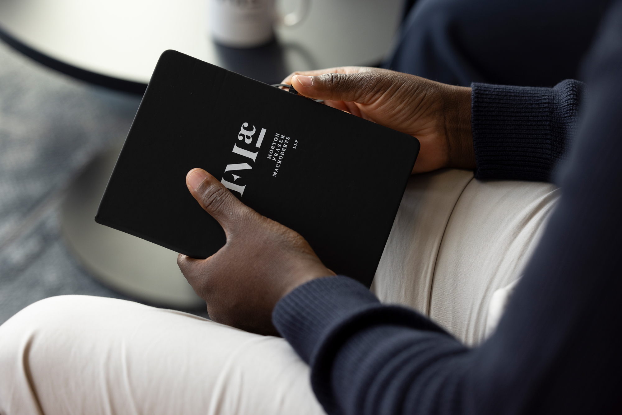 Hands holding a black notebook with white lettering, viewed from above while seated indoors