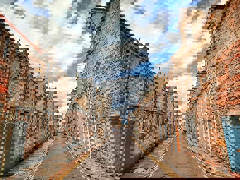 Historic Scottish street lined with traditional stone buildings, viewed from the centre of the road under a cloudy sky