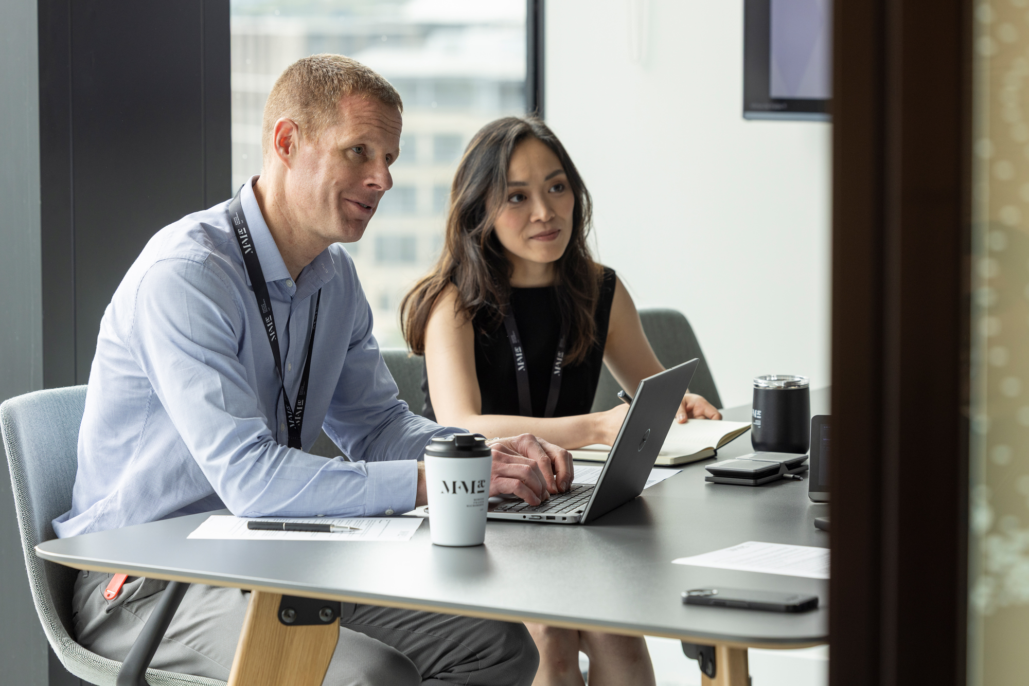 Two colleagues working together at a desk in a modern office, reviewing work on a laptop with documents and a coffee cup nearby.