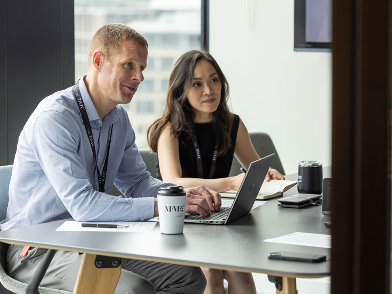 Two colleagues working together at a desk in a modern office, reviewing work on a laptop with documents and a coffee cup nearby.